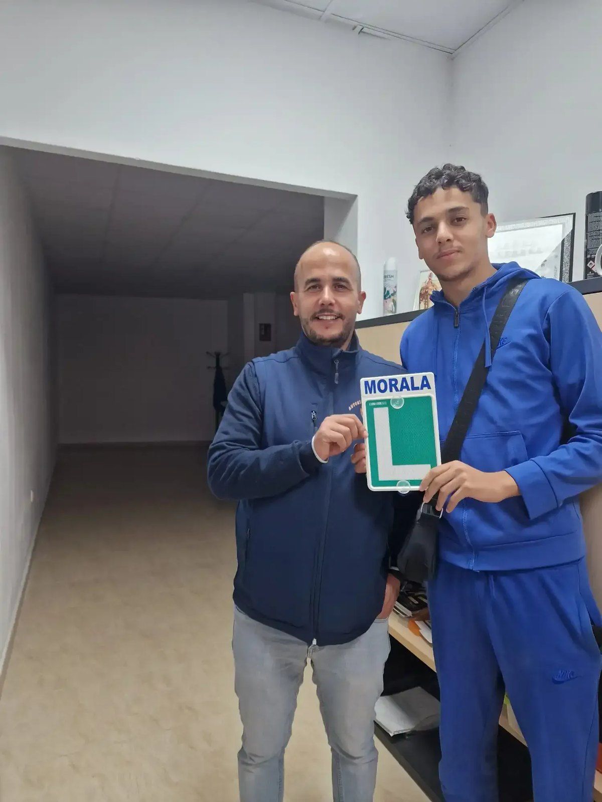 Alumno aprobado sujetando la placa L de Morala con su profesor, dentro de la autoescuela